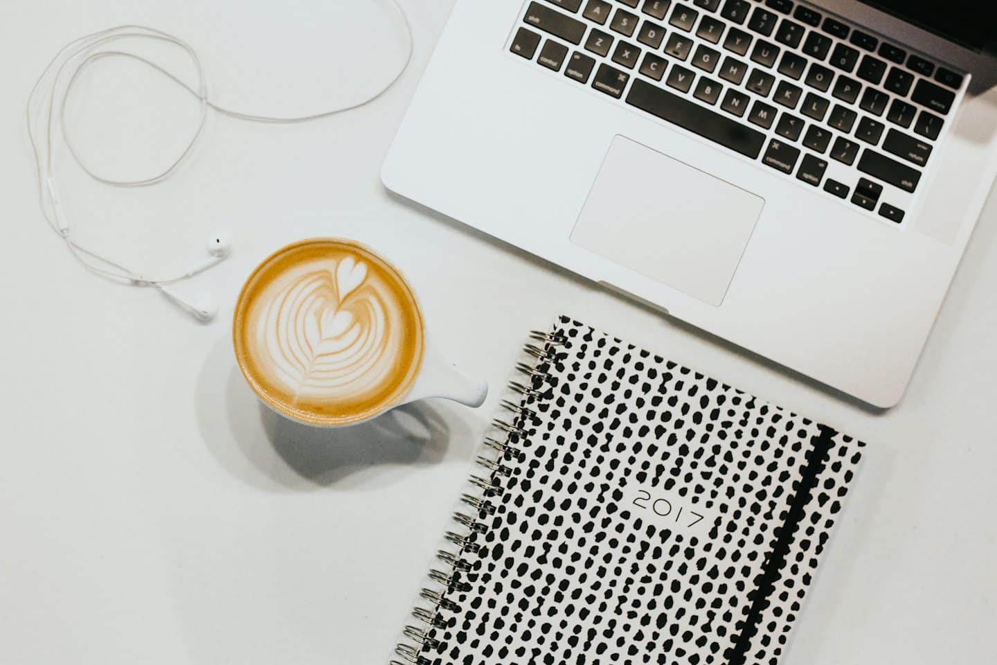 Laptop, tazza di caffè, auricolari e agenda su una scrivania bianca.